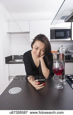 Stock Photo - Happy young woman reading text message on smart phone in kitchen. Fotosearch