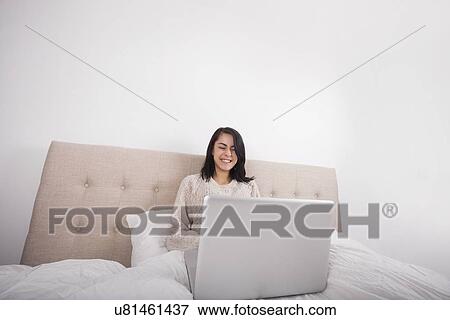 Happy young woman using laptop on bed View Large Photo Image Stock Photo - Happy young woman using laptop on bed. Fotosearch