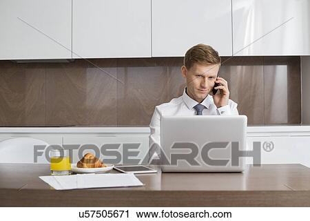 Mid adult businessman on call while using laptop at breakfast table View Large Photo Image Stock Image - Mid adult businessman on call while using laptop at breakfast table. Fotosearch
