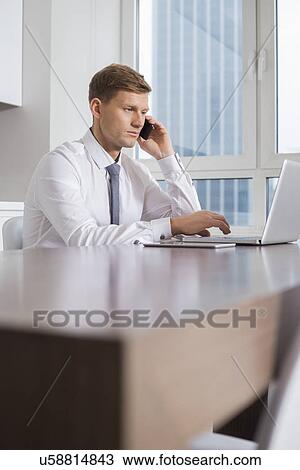 Stock Image - Mid adult businessman on call while using laptop at home. Fotosearch