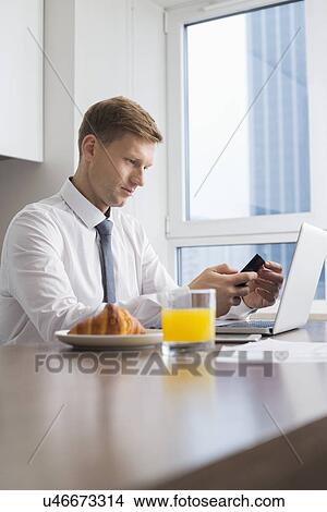Picture - Mid adult businessman using cell phone with laptop on breakfast table. Fotosearch