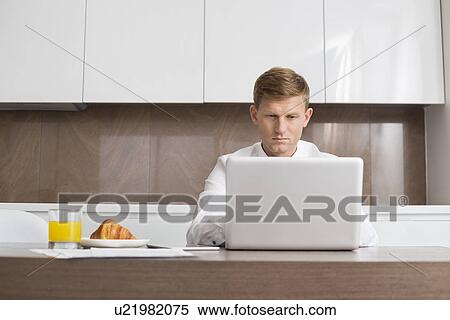 Stock Photography - Mid adult businessman using laptop at breakfast table. Fotosearch