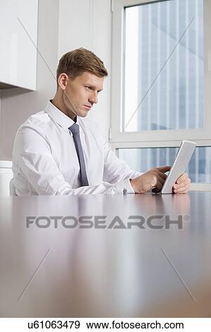 Stock Photo - Mid adult businessman using tablet computer at kitchen table. Fotosearch