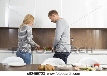 Stock Image - Mid adult couple preparing meal together in kitchen. Fotosearch