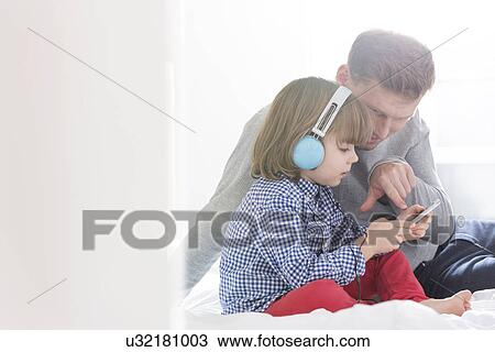 Stock Image - Mid adult father with boy listening music on headphones in bedroom. Fotosearch