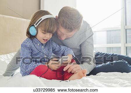Stock Photography - Mid adult father with boy listening music on headphones in bedroom. Fotosearch