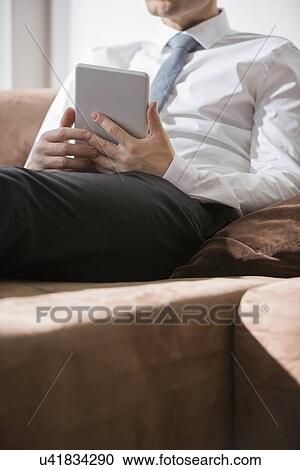 Stock Image - Midsection of businessman holding digital tablet on sofa at home. Fotosearch