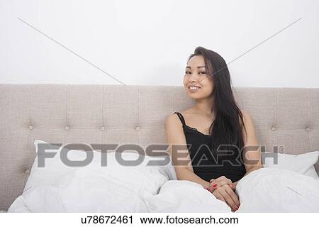 Stock Image - Portrait of beautiful young woman in nightwear sitting on bed. Fotosearch