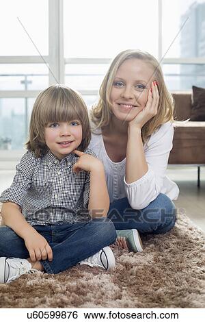 Stock Photograph - Portrait of happy mother sitting with boy on rug in living room. Fotosearch