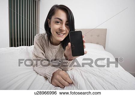 Portrait of happy woman showing smart phone while lying in bed View Large Photo Image Stock Photograph - Portrait of happy woman showing smart phone while lying in bed. Fotosearch