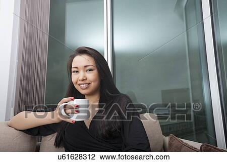 Portrait of happy young woman holding coffee mug in living room View Large Photo Image Stock Image - Portrait of happy young woman holding coffee mug in living room. Fotosearch