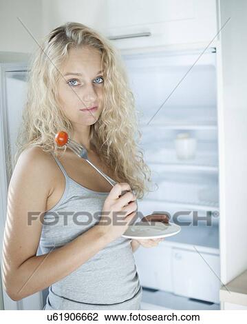 Stock Image - Portrait of sad woman having cherry tomato in kitchen. Fotosearch