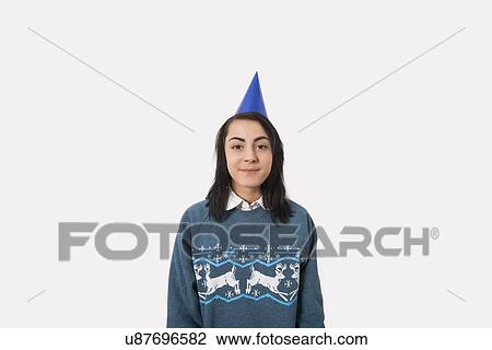Stock Image - Portrait of woman wearing Christmas jumper and party hat against gray background. Fotosearch