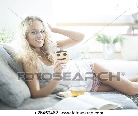 Stock Image - Portrait of young woman having cookie while relaxing on sofa. Fotosearch