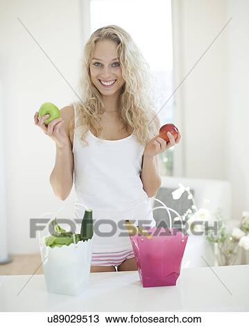 Stock Image - Portrait of young woman holding apples at kitchen counter. Fotosearch