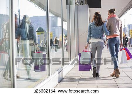Picture - Rear view of young female friends with shopping bags walking on sidewalk. Fotosearch