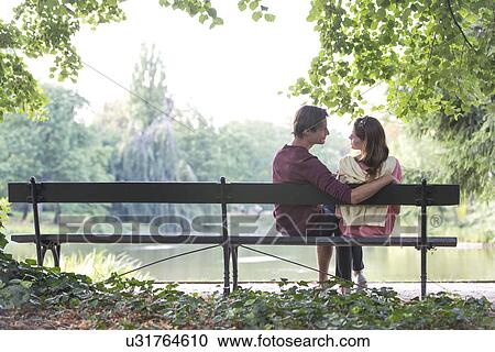 Stock Image - Romantic young couple sitting on park bench by lake. Fotosearch