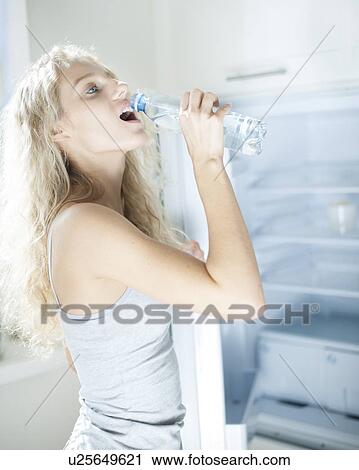 Stock Image - Side view of young woman drinking water from bottle by refrigerator. Fotosearch