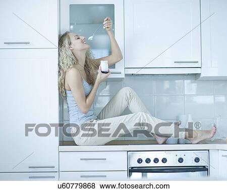 Stock Photo - Side view of young woman eating yogurt while sitting on kitchen counter. Fotosearch