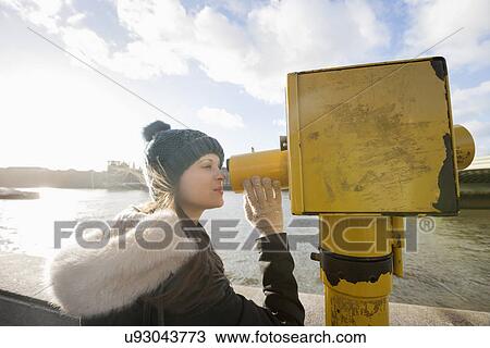 Stock Image - Side view of young woman in hooded sweatshirt looking through telescope by river Thames, London, UK. Fotosearch