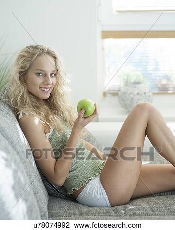 Stock Image - Side view portrait of young woman holding apple while sitting on sofa in house. Fotosearch