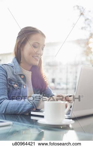 Stock Image - Smiling young woman using laptop at sidewalk cafe. Fotosearch