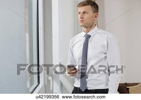 Stock Photograph - Thoughtful businessman holding smart phone while looking through window at home. Fotosearch