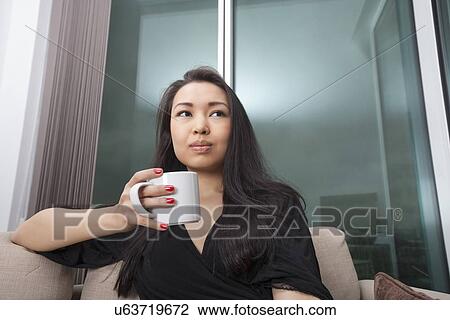 Thoughtful young woman having coffee in living room View Large Photo Image Stock Image - Thoughtful young woman having coffee in living room. Fotosearch
