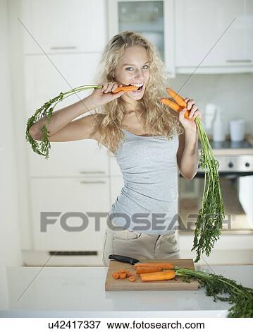 Stock Photo - Woman eating carrot at kitchen counter. Fotosearch