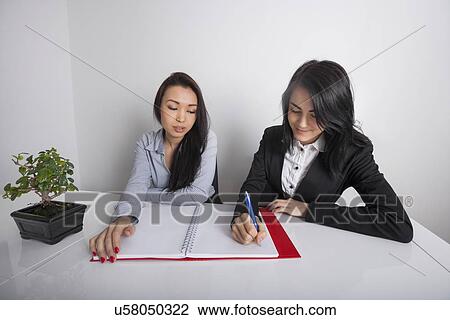 Young businesswoman with colleague writing in spiral notebook at office desk View Large Photo Image Stock Image - Young businesswoman with colleague writing in spiral notebook at office desk. Fotosearch