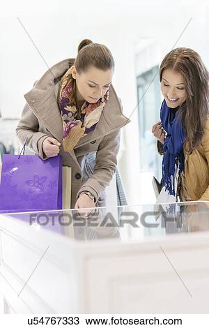 Stock Image - Young female friends looking at display cabinet in store. Fotosearch