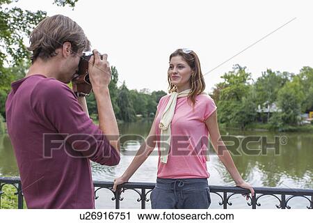 Stock Image - Young man photographing woman at lakeside. Fotosearch