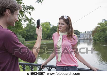 Stock Photo - Young man photographing woman through cell phone at lakeside. Fotosearch