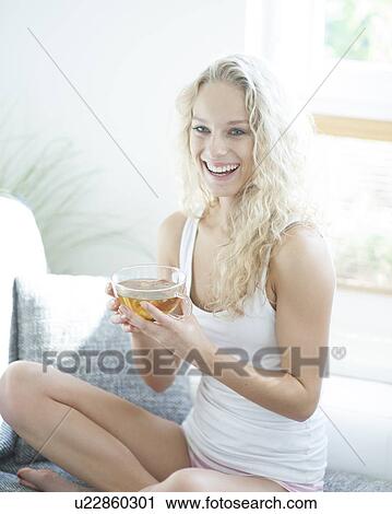 Stock Image - Young woman holding herbal tea cup while sitting on sofa. Fotosearch