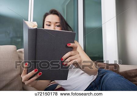 Stock Photo - Young woman reading book in living room. Fotosearch