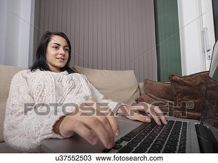 Stock Image - Young woman using laptop in living room. Fotosearch
