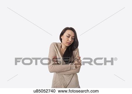 Stock Image - Young woman with arms crossed shivering against gray background. Fotosearch