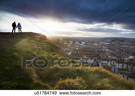 Stock Photo - A young couple hold hands in front of the view of the City of Edinburgh from Salisbury Crags. Fotosearch