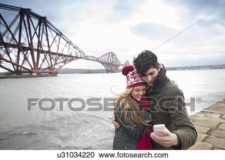 Stock Image - A young couple pose in front of the Forth Rail Bridge in Queensferry, near Edinburgh, Scotland. Fotosearch