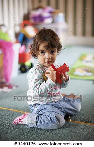 Stock Photograph - Female toddler sitting on playroom floor playing with a red toy. Fotosearch