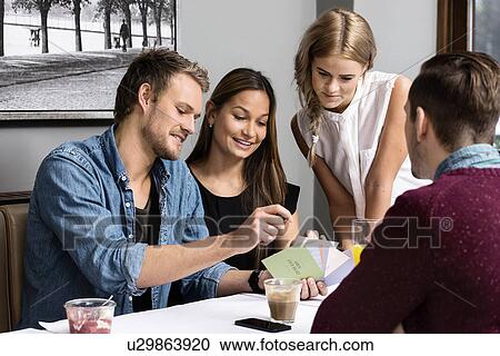 Stock Image - Four young adult friends sitting in cafe looking at cards. Fotosearch