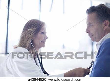 Stock Image - Mature female doctor holding stethoscope to patients chest. Fotosearch