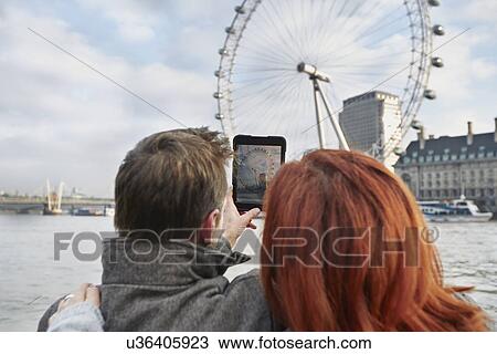 Stock Image - Mature tourist couple photographing London Eye, London, UK. Fotosearch