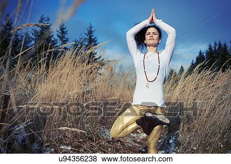 Stock Photo - Mid adult woman doing standing tree yoga pose in forest. Fotosearch