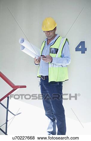 Stock Photo - Senior male project manager holding blueprints and texting. Fotosearch