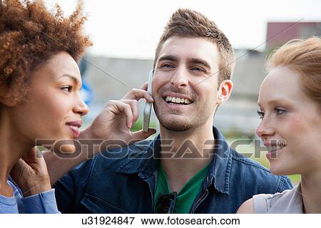 Stock Photo - Three young adult friends, one talking on smartphone. Fotosearch