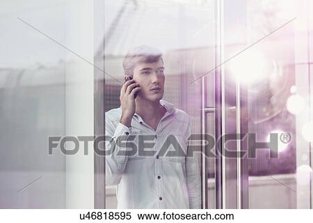 Stock Photography - Young man behind illuminated reflective glass talking on smartphone. Fotosearch