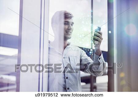 Stock Image - Young man behind reflective glass looking at illuminated smartphone. Fotosearch