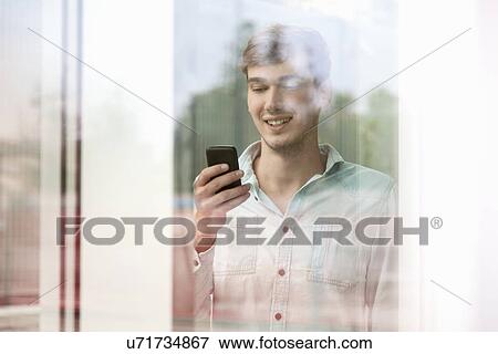 Stock Photo - Young man behind reflective glass looking at smartphone. Fotosearch