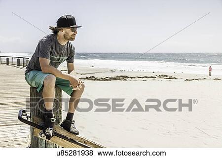 Stock Photo - Young man sitting on wooden post, Laguna Beach, California, USA. Fotosearch
